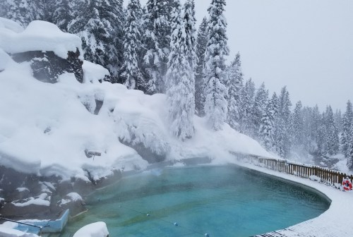 A steaming hot spring surrounded by snow-covered trees and rocky terrain in a winter landscape.