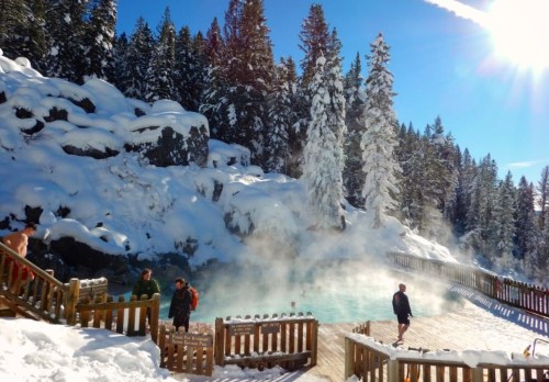 A snowy landscape with a steaming hot spring, surrounded by tall trees and people enjoying the warm water.