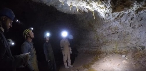 A group of people wearing helmets with lights stands in a dimly lit cave, observing the rocky surroundings.