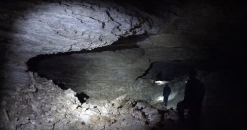 A dark cave interior with rocky walls, illuminated by the beams of two flashlights held by figures in the distance.