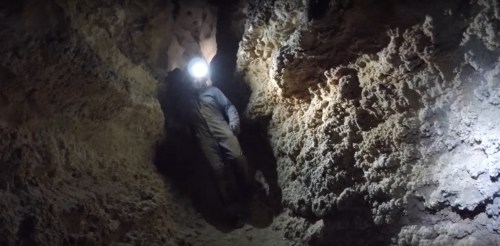 A miner wearing a headlamp explores a rocky cave, surrounded by textured walls and dim lighting.