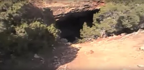A rocky landscape with a cave entrance surrounded by shrubs and dirt.