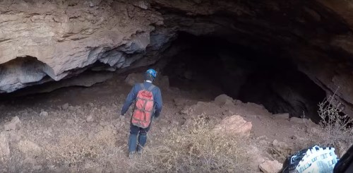 A person in a helmet and backpack stands at the entrance of a rocky cave, surrounded by dirt and sparse vegetation.