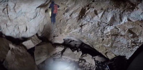 A person in climbing gear navigates a rocky cave, illuminated by a flashlight, with large boulders scattered below.