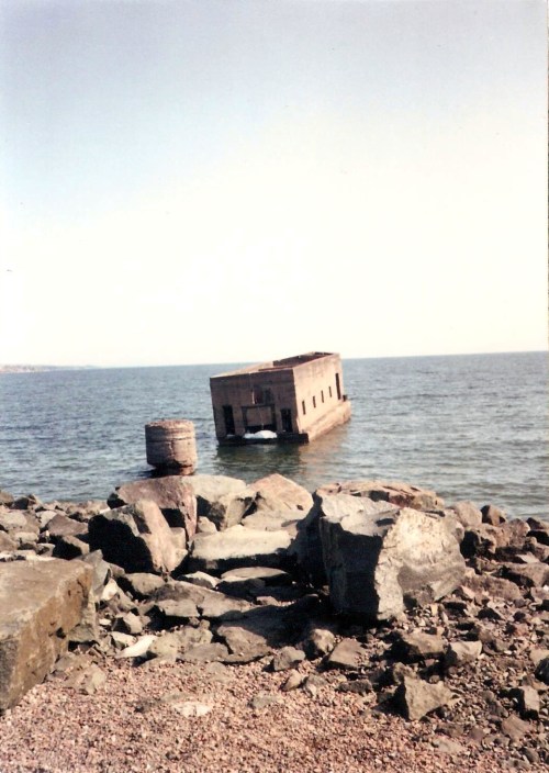 A partially submerged structure on a rocky shoreline, with calm water and a clear sky in the background.
