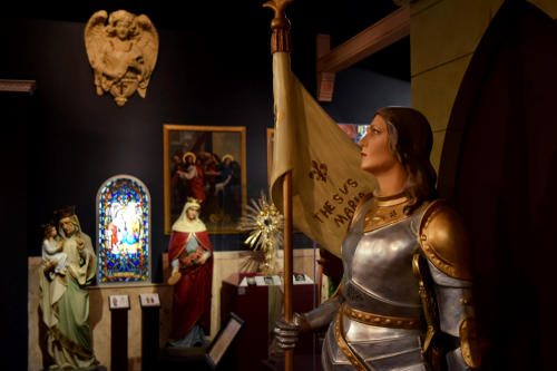 A museum display featuring a knight statue holding a flag, surrounded by religious art and stained glass.