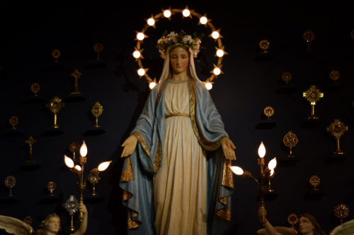 A statue of the Virgin Mary with a floral crown, surrounded by religious symbols and candles in a dimly lit setting.