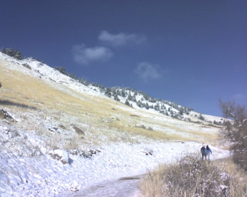 Two people walk along a snowy path beside a grassy hillside under a clear blue sky.