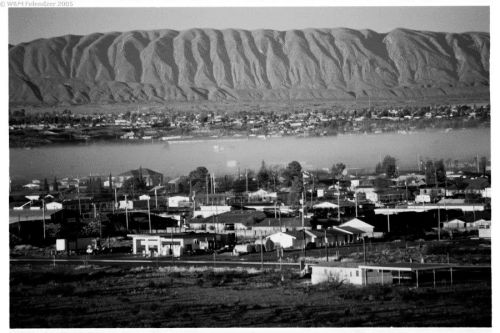 Black and white landscape of a small town with mountains in the background and a river reflecting the scenery.