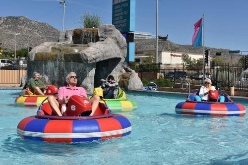 Four people in colorful bumper boats enjoying a sunny day in a pool with a rock waterfall in the background.
