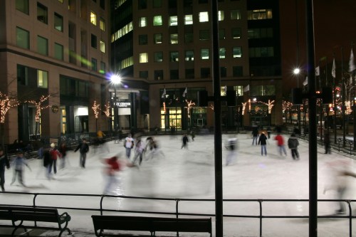 A bustling outdoor ice skating rink at night, with skaters in motion and city buildings illuminated in the background.