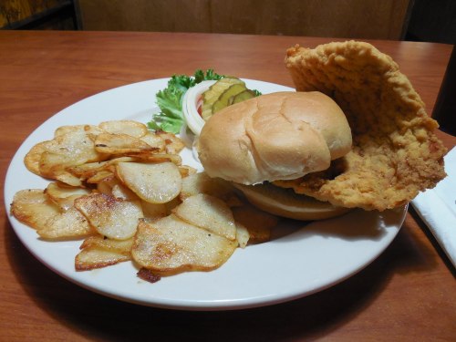 A plate with a fried chicken sandwich, lettuce, pickles, and a side of crispy potato chips.