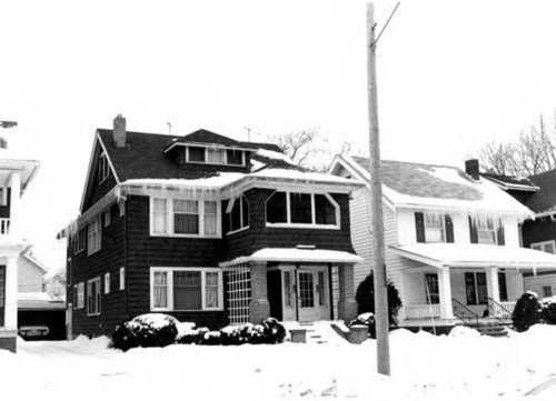 Two large houses covered in snow, with a power pole in the foreground and trees in the background. Black and white photo.
