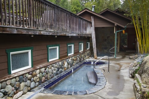 A serene outdoor spa area featuring a stone-lined hot tub beside a wooden deck and bamboo plants.