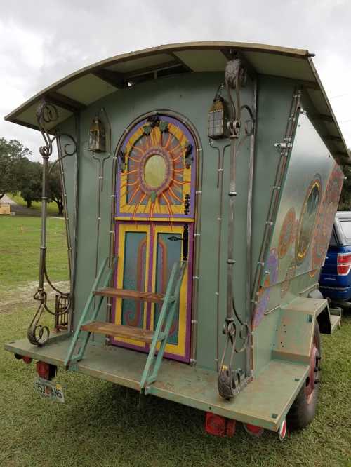 Colorful vintage caravan with a decorative door and steps, set in a grassy area under a cloudy sky.