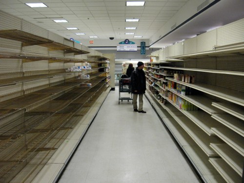 A grocery store aisle with mostly empty shelves and two shoppers with a cart, indicating low stock or a supply shortage.