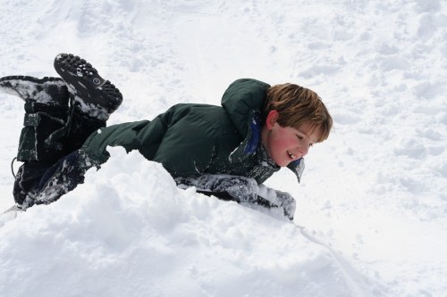 A young boy in a green jacket plays in the snow, smiling as he slides down a snowy hill.