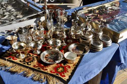 A display of various silverware and glassware arranged on a blue tablecloth with a decorative rug underneath.