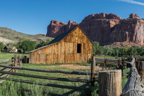 A rustic wooden barn surrounded by green fields and rocky cliffs under a clear blue sky.