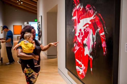 A woman holds a child while pointing at a vibrant artwork in a gallery.