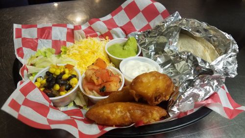 A plate with a foil-wrapped tortilla, salad, cheese, guacamole, sour cream, and crispy fried items on a checkered paper.