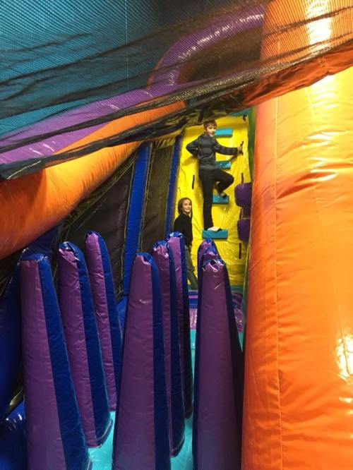 Two children play on a colorful indoor inflatable obstacle course with slides and climbing walls.