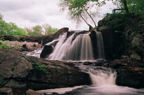 A serene waterfall cascades over rocky terrain, surrounded by lush greenery under a cloudy sky.