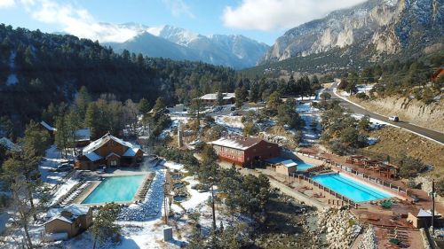 Aerial view of a snowy resort with pools, surrounded by mountains and trees under a clear blue sky.