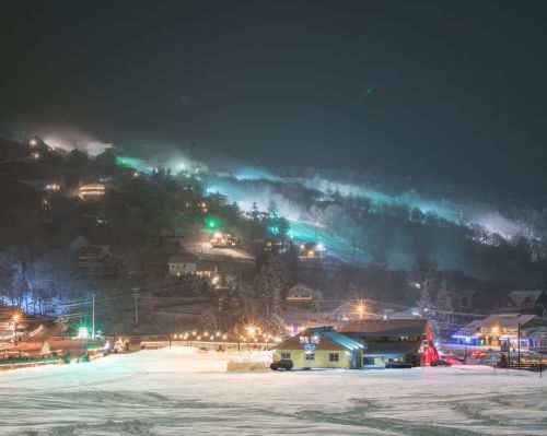 A snowy landscape at night, with colorful lights on a hill and houses illuminated in the distance.