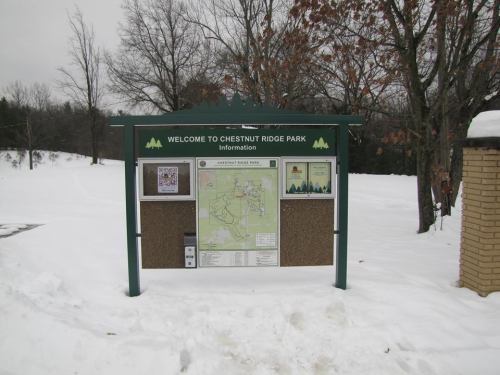 Sign at Chestnut Ridge Park displaying a map and information, surrounded by snow-covered trees.