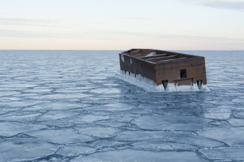 A weathered wooden structure floats on a frozen sea, surrounded by cracked ice under a pale sky.