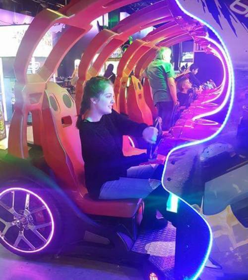 A person sits in a colorful arcade racing game, gripping the steering wheel, with vibrant lights surrounding them.