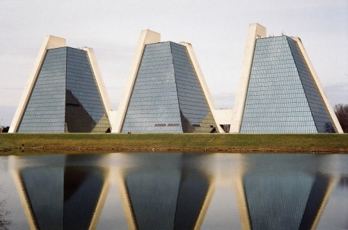 Three modern triangular glass buildings reflected in a calm body of water, surrounded by green grass and trees.