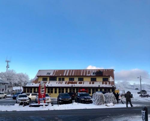 A snowy scene featuring a yellow building with a brown roof, surrounded by parked cars and a clear blue sky.