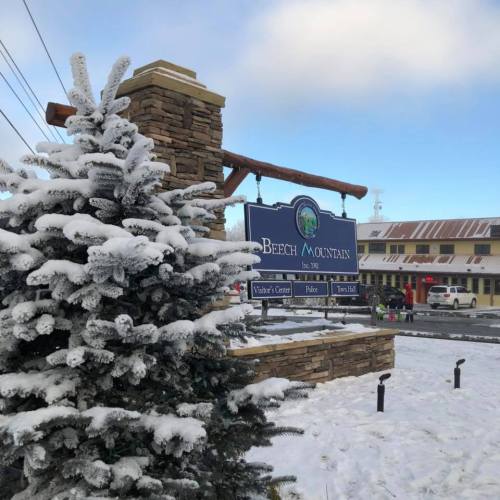 A snowy landscape featuring a sign for Beech Mountain, surrounded by frosted trees and a wintery scene.
