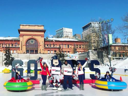 A group of cheerleaders and a mascot pose on an ice rink with large "GO BOLTS" letters in the background.