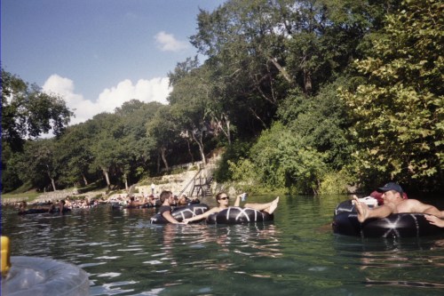 People relaxing on inner tubes in a river, surrounded by trees and a sunny sky.