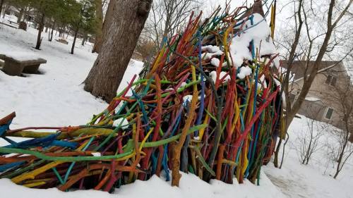 Colorful sticks arranged in a whimsical structure, partially covered in snow, set in a winter park landscape.