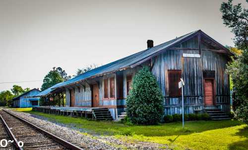 A weathered wooden train station beside railway tracks, surrounded by greenery and a clear sky.