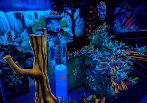 A vibrant, neon-lit mini-golf course featuring a robot, trees, and a waterfall backdrop in a jungle setting.