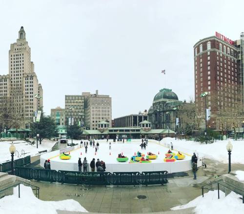 A winter scene featuring an ice rink with people skating and colorful bumper cars, surrounded by snowy buildings.