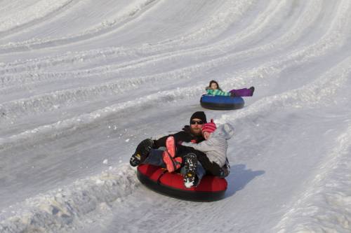 Two people sledding down a snowy hill on inner tubes, with one adult holding a child in front.