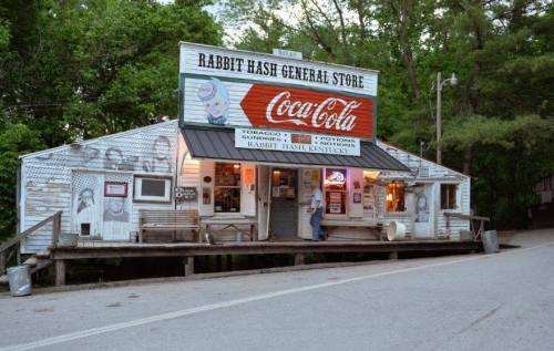 A rustic general store with a Coca-Cola sign, surrounded by trees, featuring a porch and vintage decor.