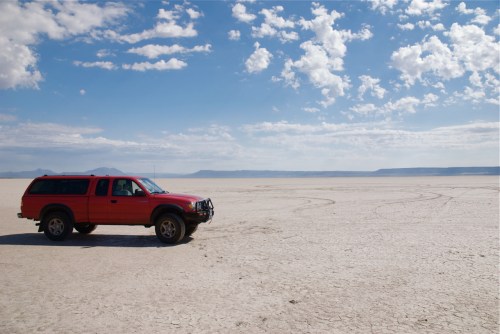 A red SUV parked on a dry, cracked landscape under a blue sky with scattered clouds.
