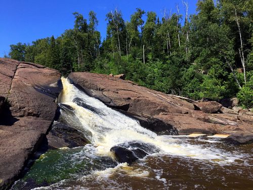 A waterfall cascading over rocky terrain, surrounded by lush green trees under a clear blue sky.