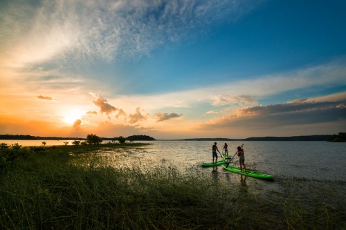 Three people paddleboarding on a calm lake at sunset, with vibrant clouds and lush greenery in the foreground.