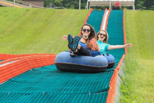 Two women joyfully tubing down a grassy slope on a summer day, wearing sunglasses and smiling.