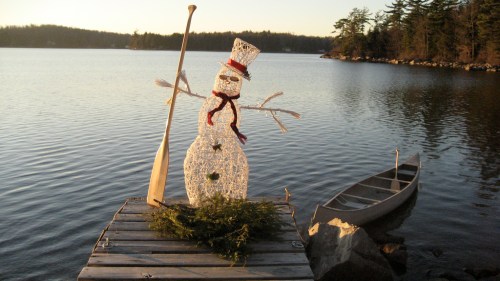 A snowman with a hat and scarf stands on a dock by a calm lake, with a canoe nearby and trees in the background.