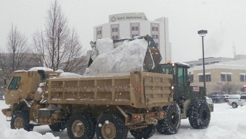 A military truck and loader clear heavy snow in a city during a snowstorm, with a hospital in the background.