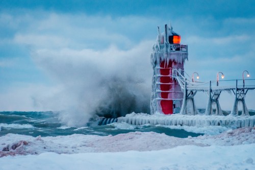 A frozen lighthouse stands against crashing waves, surrounded by ice and snow under a cloudy sky.